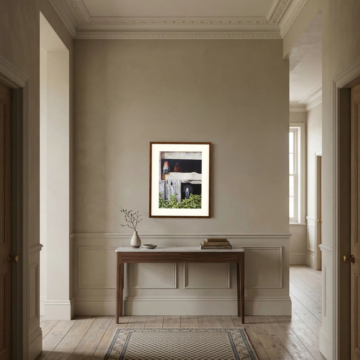 A framed (dark oak frame) art paper print with a freja passepartout depicting three horses peeking out from a rustic wooden stable, with green foliage in the foreground, hangs on a beige wall in a classic hallway setting above a dark wood console table. 