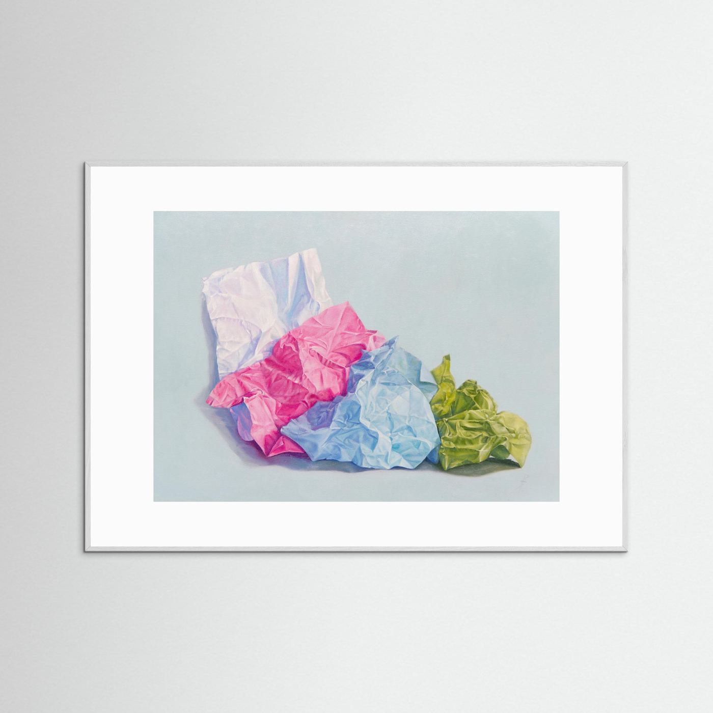 A framed (white frame) paper art print with a white passepartout featuring a still life painting of crumpled colored paper in pink, blue, white, and green on a light blue-gray background, displayed against a neutral light gray wall.