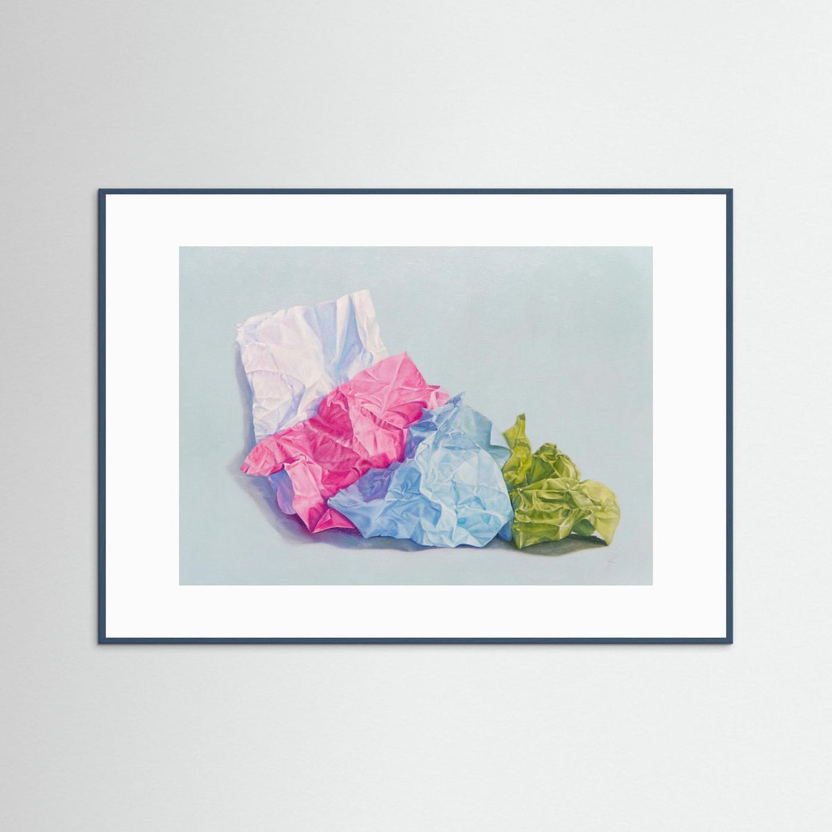 A framed (teal wood) with a white passepartout paper art print featuring a still life painting of crumpled colored paper in pink, blue, green, and white on a light blue-gray background, displayed against a neutral light gray wall.