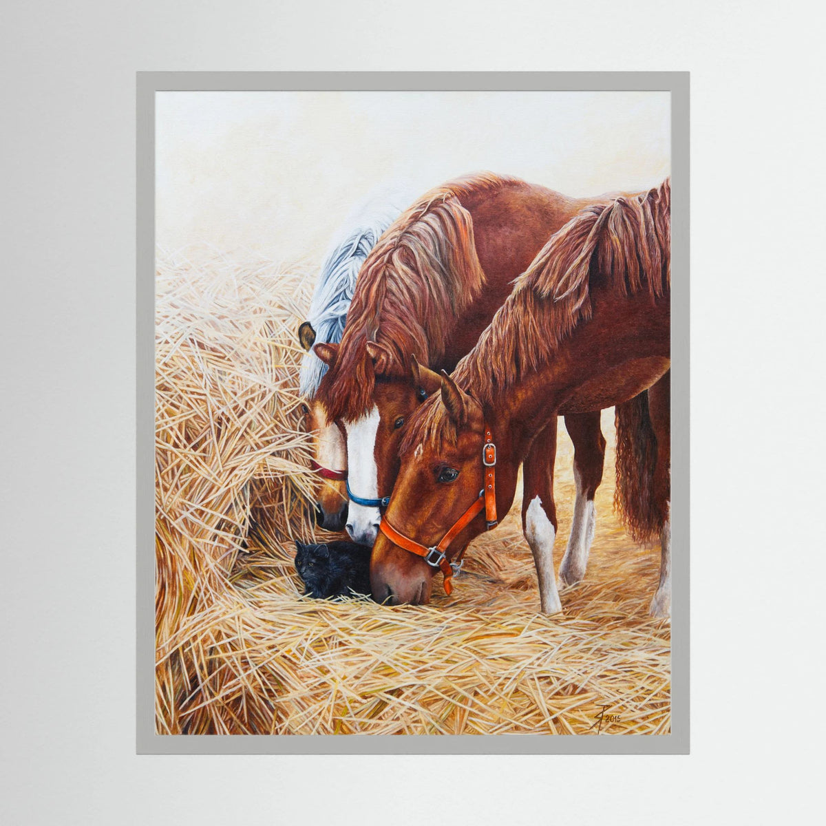 A framed (white frame) paper art print depicting three young horses on a bed of golden straw, with a small black cat nestled among the straw. The print is presented with a light gray frame against a neutral light gray background.