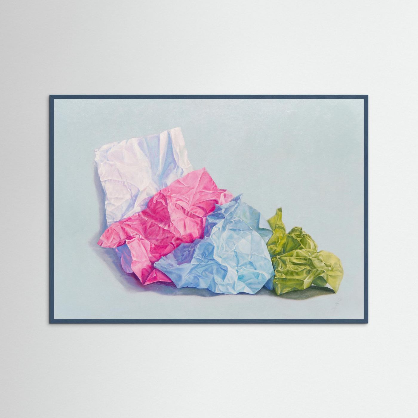A framed (teal wood) paper art print depicting crumpled colored paper in pink, blue, green, and white against a pale blue-gray background, displayed on a light gray wall.