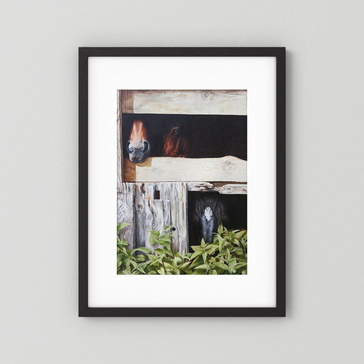 A framed (black frame) paper art print with a white passepartout featuring a rustic barn scene with horses peeking out from stalls, mounted against a light gray wall.