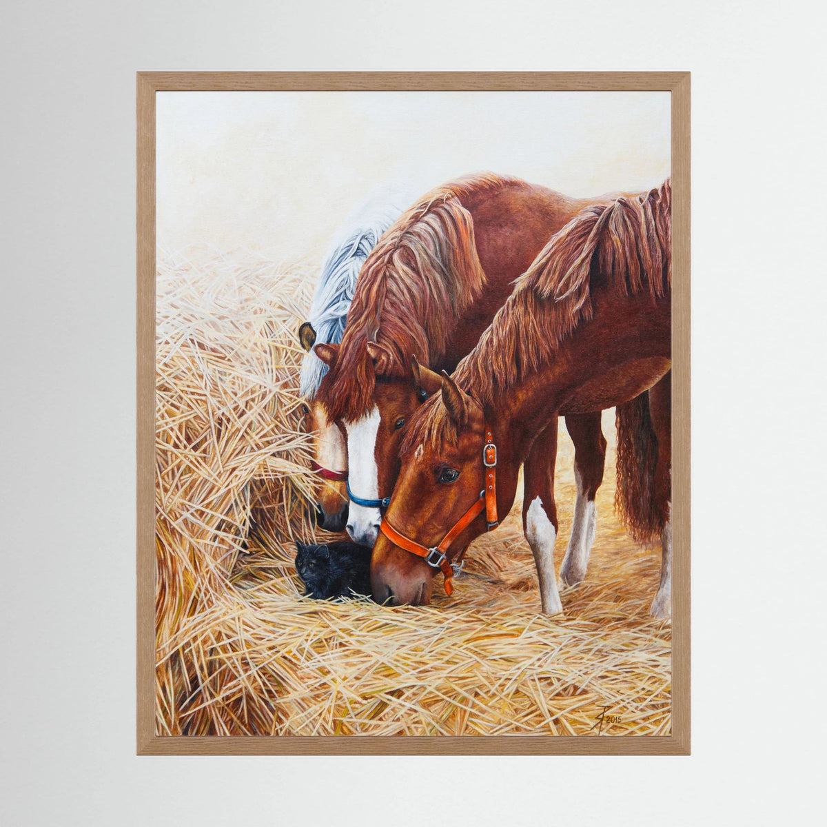 A framed (nature oak) paper art print depicting three young horses with a small black cat nestled in the straw displayed against a neutral light gray background.