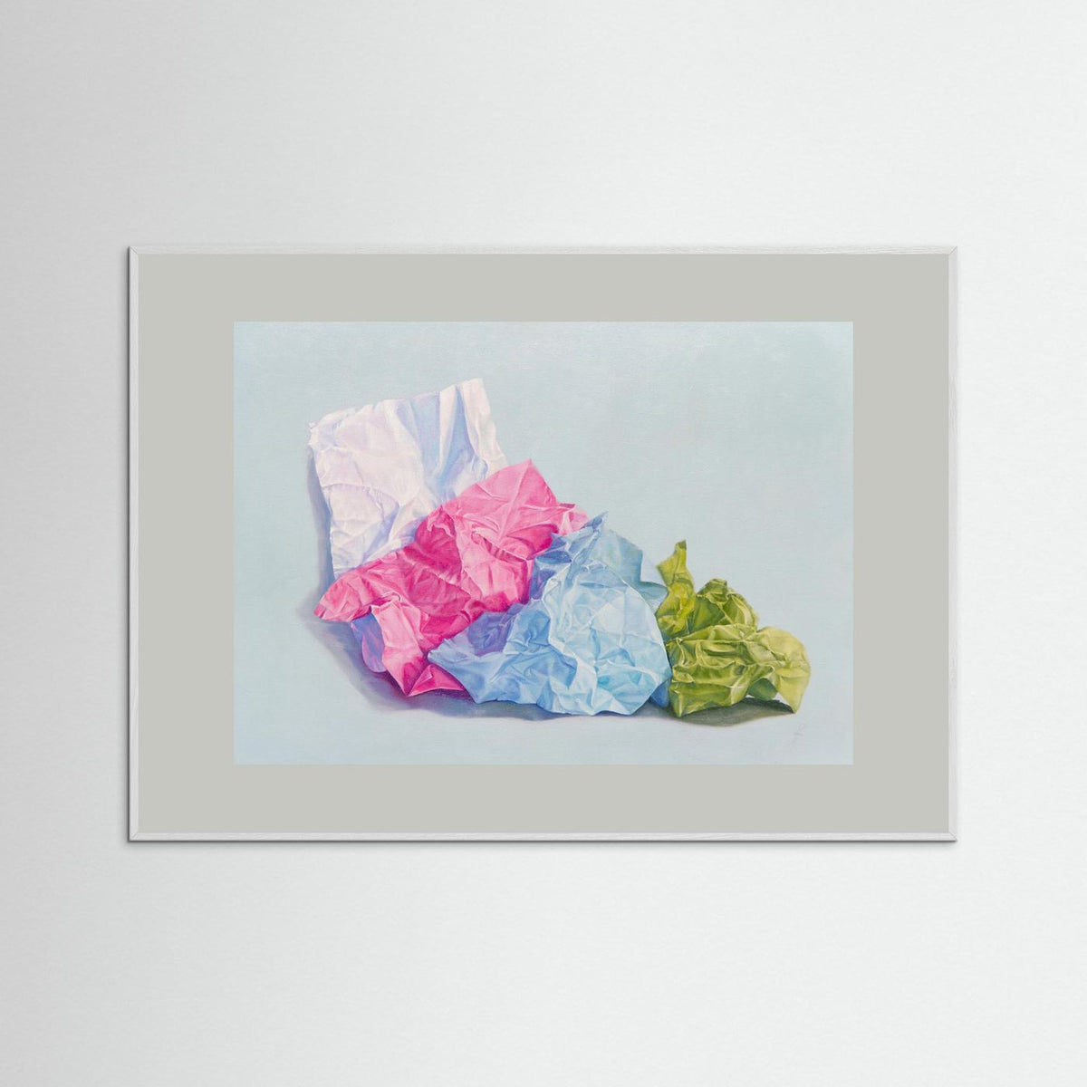 A framed (white frame) paper art print with a grey passepartout featuring a still life painting of crumpled colored paper in pink, blue, white, and green on a pale blue background, displayed against a light gray wall.