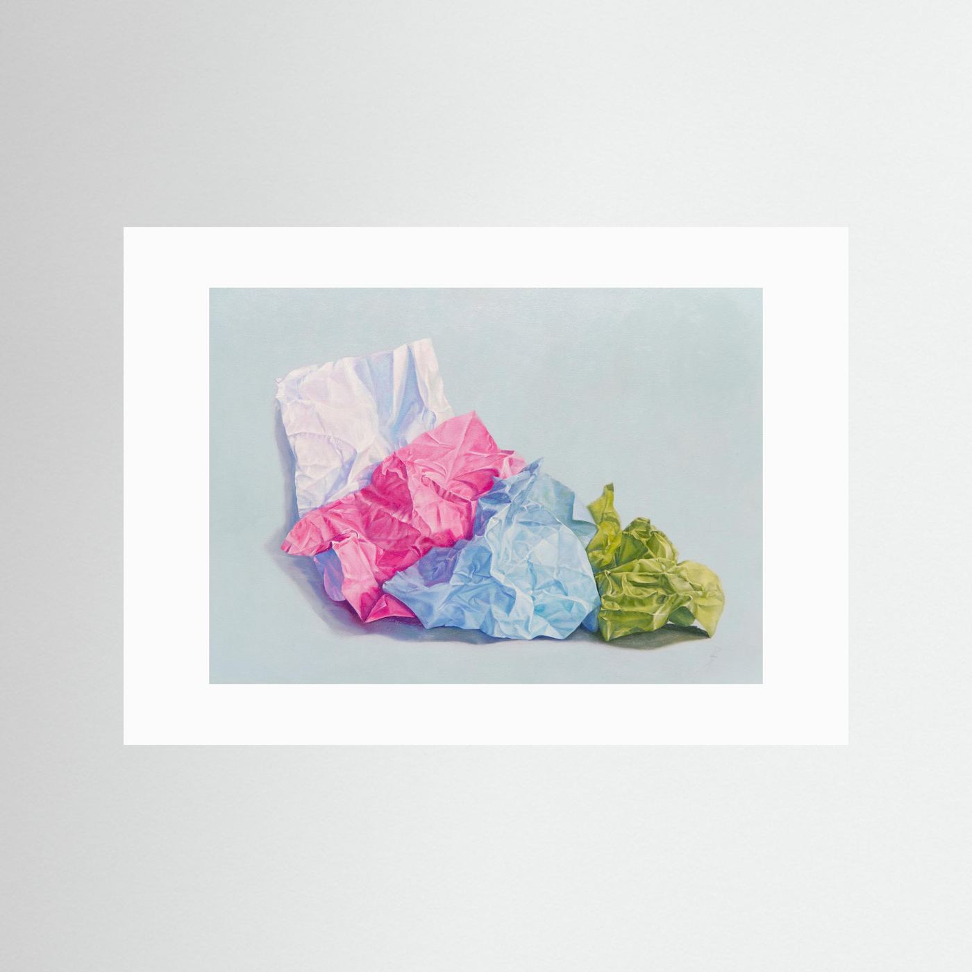 A paper art print with a white passepartout featuring a still life painting of crumpled paper in pink, blue, white, and green on a light blue-gray background, displayed against a neutral light gray wall.